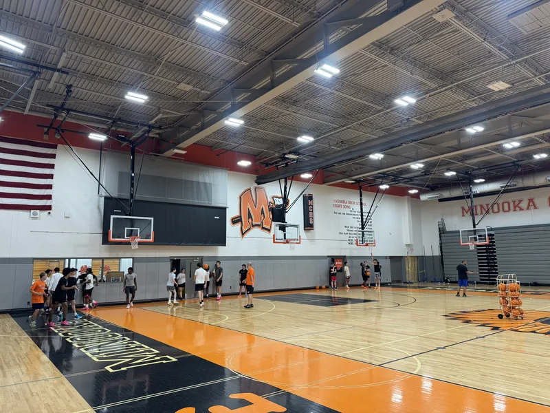 Minooka Community High School main gym in use — oversized orange "M" mascot logo, MINOOKA lettering and fight-song wall graphics above a polished orange-and-black court.