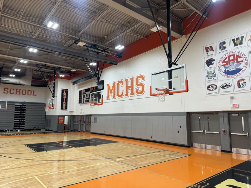 Minooka Community High School gym — dimensional orange-and-white 'MCHS' letters mounted on a white block wall beside championship banners, above a polished maple basketball court with orange-black free-throw lanes and the Southwest Prairie Conference wall panel visible.