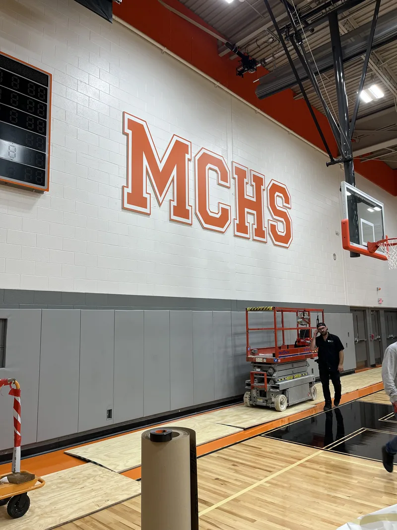 CorPro crew on a scissor lift finishing oversized orange "MCHS" letters high on the Minooka gym wall.