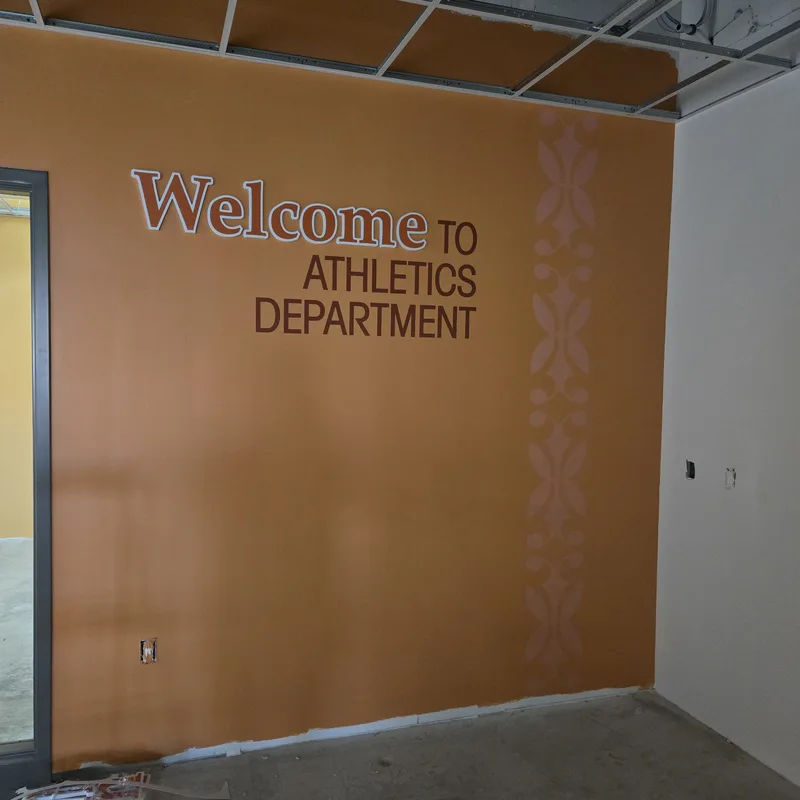 Minooka Community High School athletics office — "Welcome to Athletics Department" dimensional lettering over a burnt-orange wall graphic with a patterned vertical border.