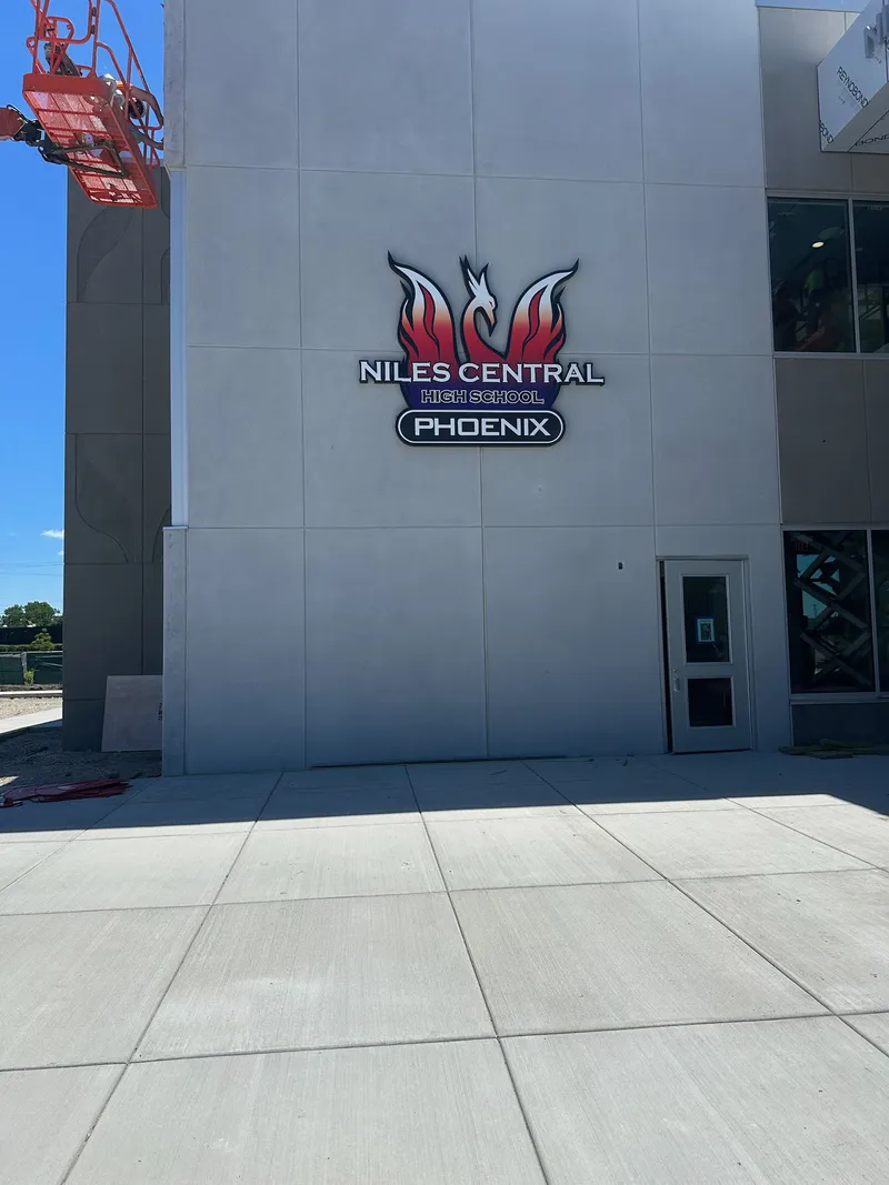 Niles Central High School Phoenix building ID — dimensional red-and-blue Phoenix mascot logo with 'NILES CENTRAL HIGH SCHOOL / PHOENIX' wordmark, mounted mid-height on a modern concrete-panel facade, install lift edge visible.