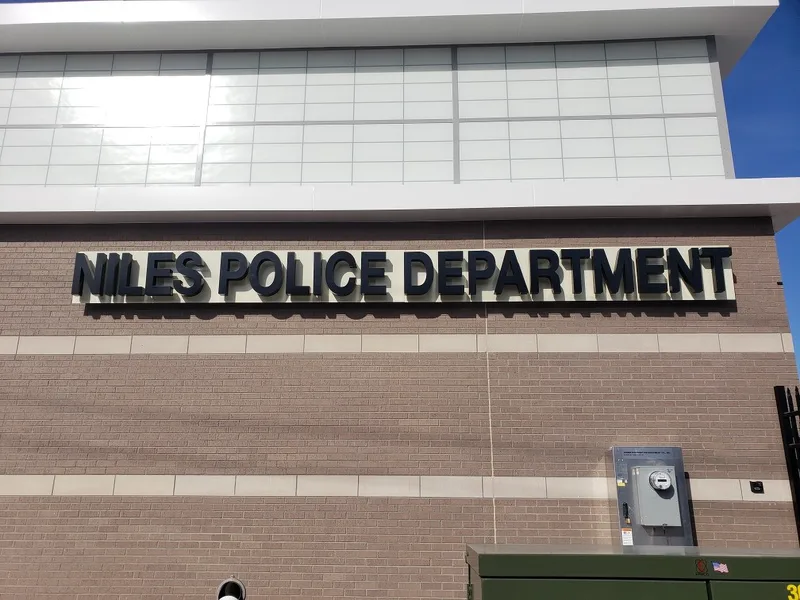 Niles Police Department — large black dimensional letters with stand-off shadow lines across a tan brick facade beneath a glass curtain wall.