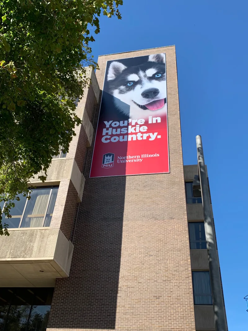 Vertical "You're in Huskie Country" banner with a close-up Huskie portrait on a NIU brick building facade.