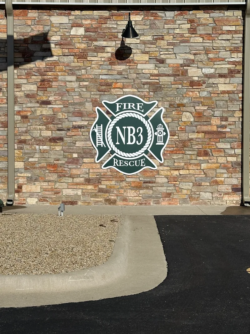 North Boone Fire District 3 station — dimensional 'NB3' fire-rescue shield emblem (axes, fire hydrant, ladder) mounted on a stacked-stone exterior wall beneath a hanging bell.