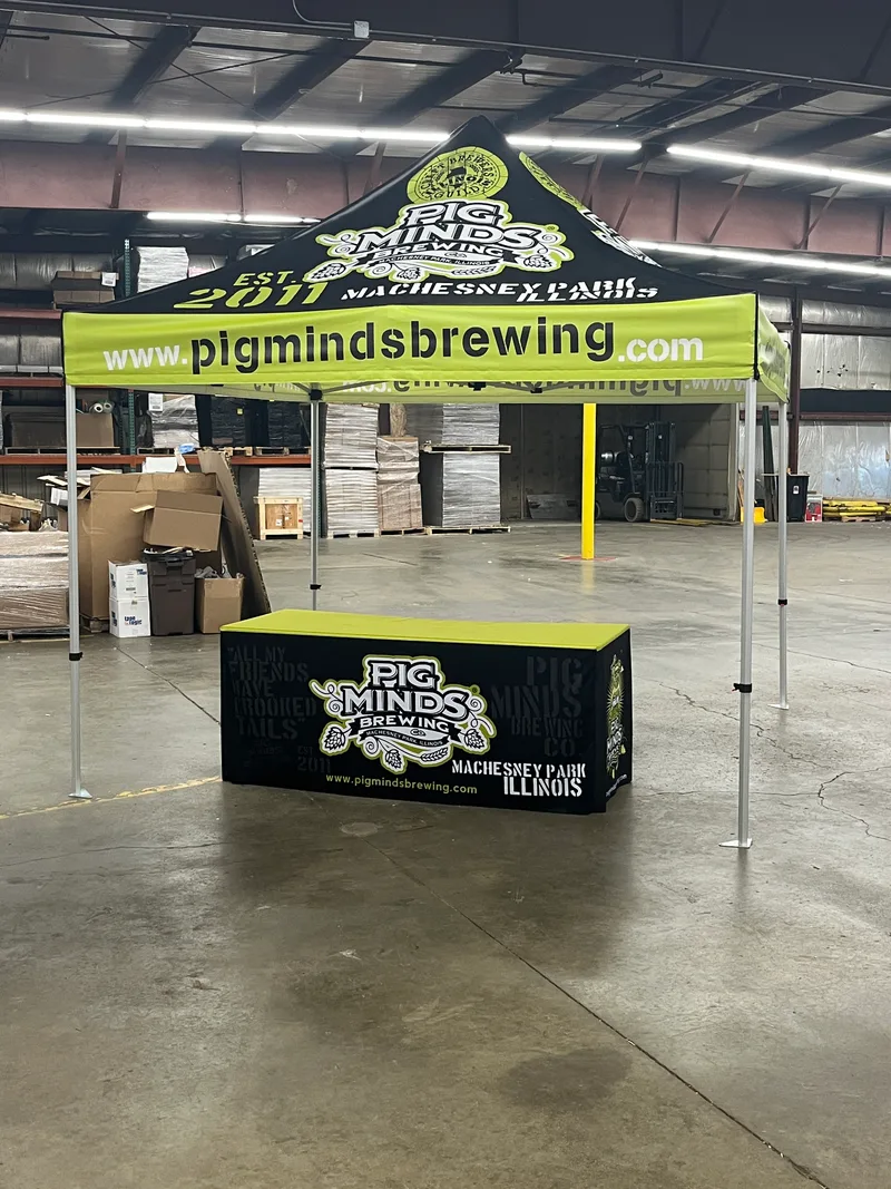 Pig Minds Brewing event kit — coordinated black-and-yellow 10x10 pop-up tent and matching black table throw with the Pig Minds skull-and-hops emblem, 'EST 2011 / Machesney Park Illinois', and pigmindsbrewing.com URL, photographed in the CorPro shop before delivery.