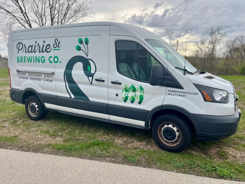 Prairie Street Brewing Co. delivery van — clean outdoor three-quarter view of a white Ford Transit with the Prairie Street peacock wordmark, 'Brewery Restaurant Events Catering Weddings & More!' tagline, and green geometric 'Cheers!' emblem, parked on grass under a cloudy spring sky.