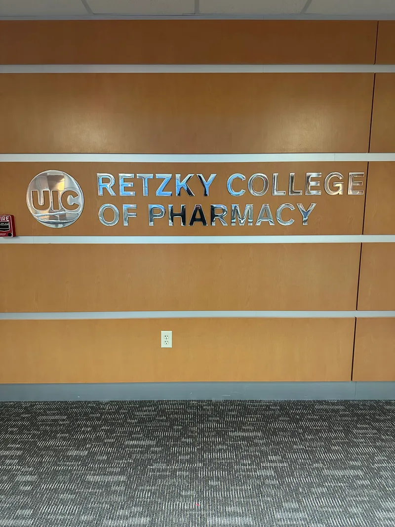 UIC Retzky College of Pharmacy — vertically-stacked brushed-metal dimensional letters with the mirrored UIC oval logo, mounted on a warm wood-laminate wall between vertical reveal lines.
