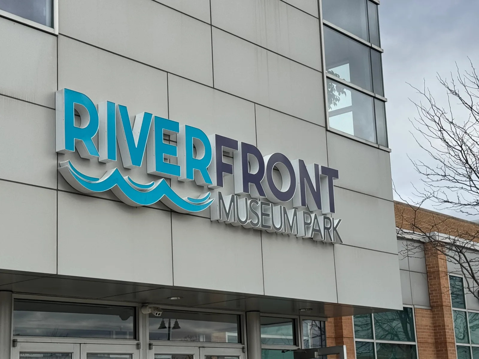 Riverfront Museum Park storefront sign — dramatic dimensional 'RIVERFRONT / MUSEUM PARK' wordmark with brushed-metal returns, animated blue wave graphic beneath 'RIVER', mounted on the modern white-panel facade above the entrance.