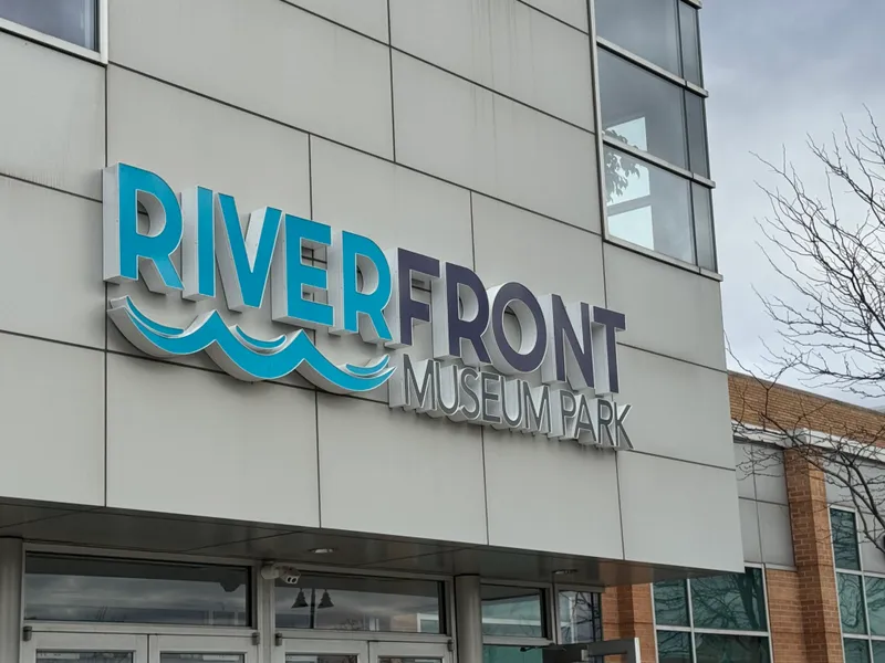 Riverfront Museum Park storefront sign — dramatic dimensional 'RIVERFRONT / MUSEUM PARK' wordmark with brushed-metal returns, animated blue wave graphic beneath 'RIVER', mounted on the modern white-panel facade above the entrance.