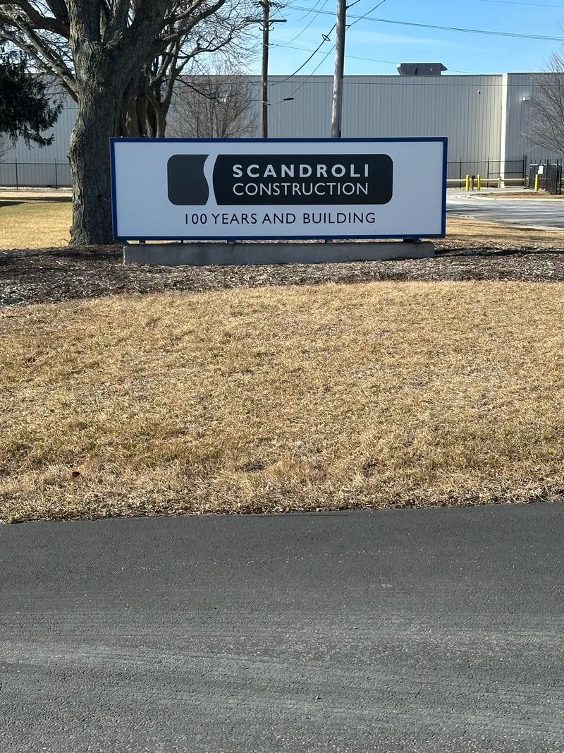 Scandroli Construction roadside identification panel — branded white-and-black panel reading 'SCANDROLI CONSTRUCTION · 100 YEARS AND BUILDING', mounted on stand-offs in front of a warehouse facade.