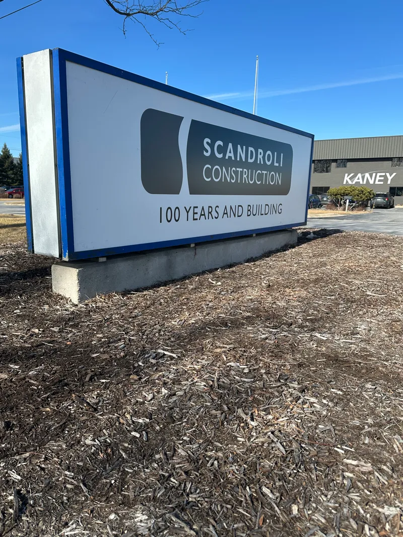 Scandroli Construction backlit cabinet sign — sleek white cabinet with dimensional 'SCANDROLI CONSTRUCTION / 100 YEARS AND BUILDING' lettering on the fascia, mounted at office-park street level with the Kaney building visible behind.