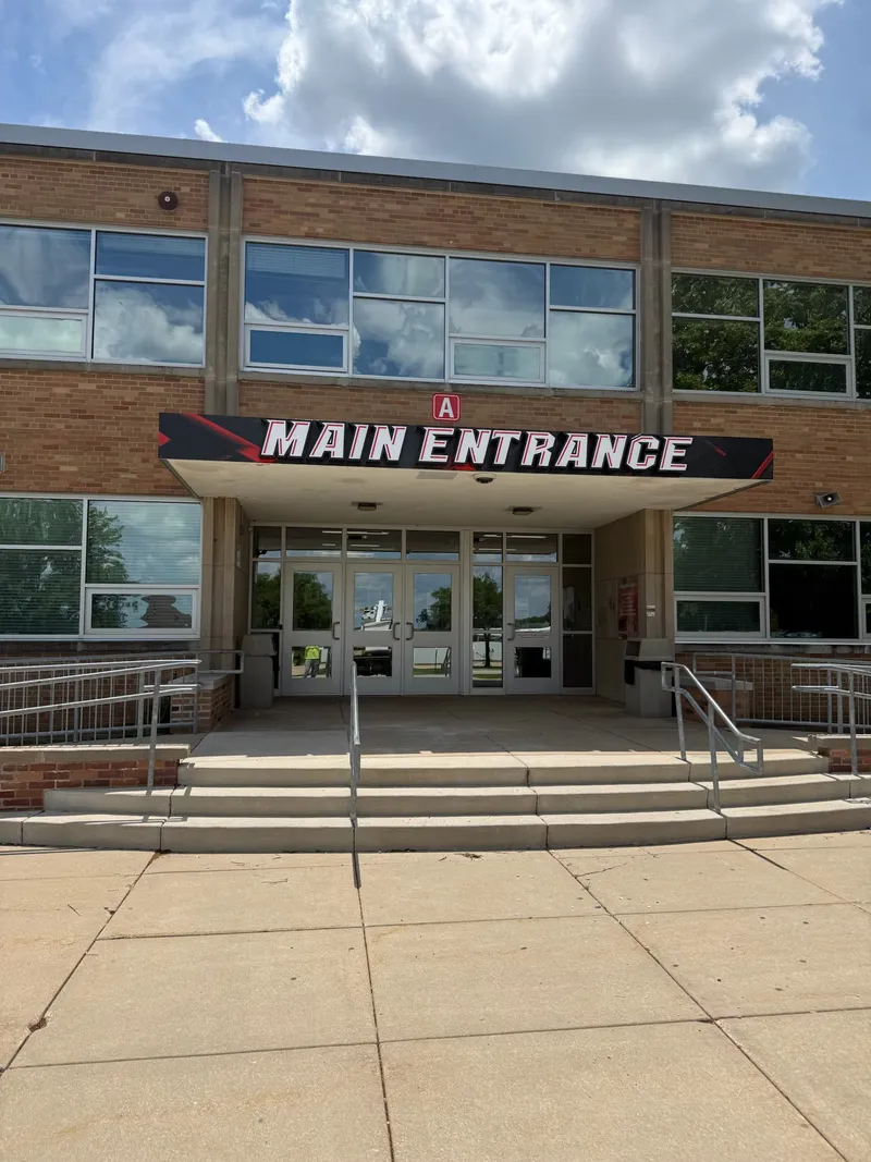 School main entrance signage — tall vertical black cabinet with dimensional 'MAIN ENTRANCE' white lettering and red athletic speed-lines graphic, mounted between two brick columns at a modern school entry.