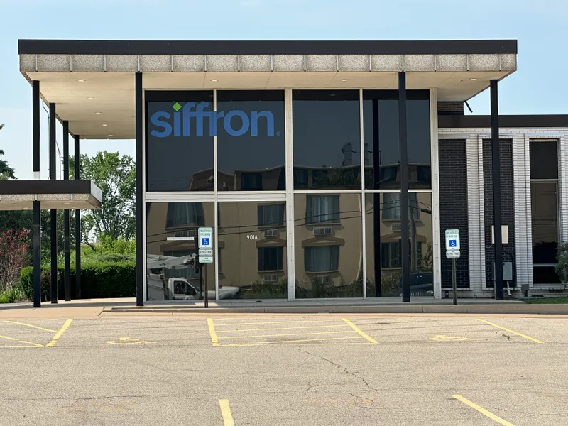 Siffron office storefront (Des Plaines, IL) — mid-century modern glass facade with dark-tinted floor-to-ceiling windows, bold blue 'siffron' dimensional window vinyl with the green diamond accent mark, aluminum framing, and concrete-and-brick wing walls.