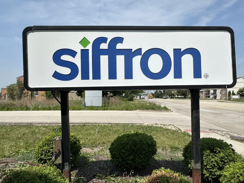Siffron roadside sign — white double-post panel with blue "siffron" logo and green diamond, set in a shrub bed along the drive.