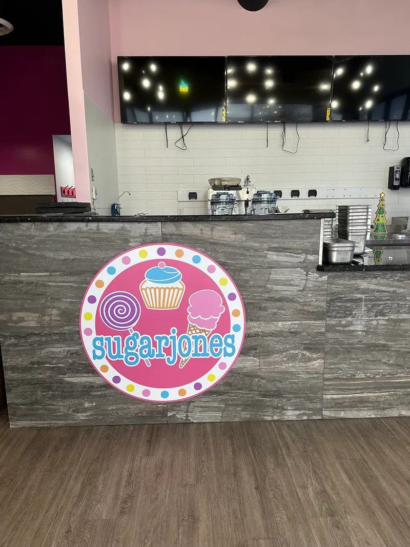 Sugar Jones ice-cream shop interior — vibrant circular 'sugarjones' dimensional logo (cupcake, ice-cream cone, swirl lollipop, polka dots) mounted on the gray-tile customer counter face beneath a row of menu TVs.
