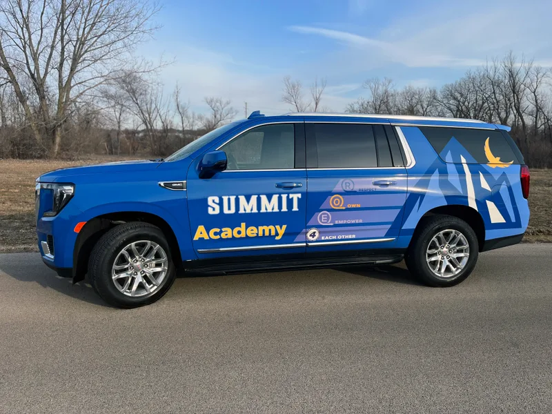 Summit Academy GMC Yukon SUV — outdoor side view of a modern blue vehicle wrap featuring 'SUMMIT Academy' wordmark in white and gold, a mountain range graphic with a golden phoenix on the rear panel, and the school's ROE4 values framework (Respect / Own / Empower / Each Other) along the rear doors.