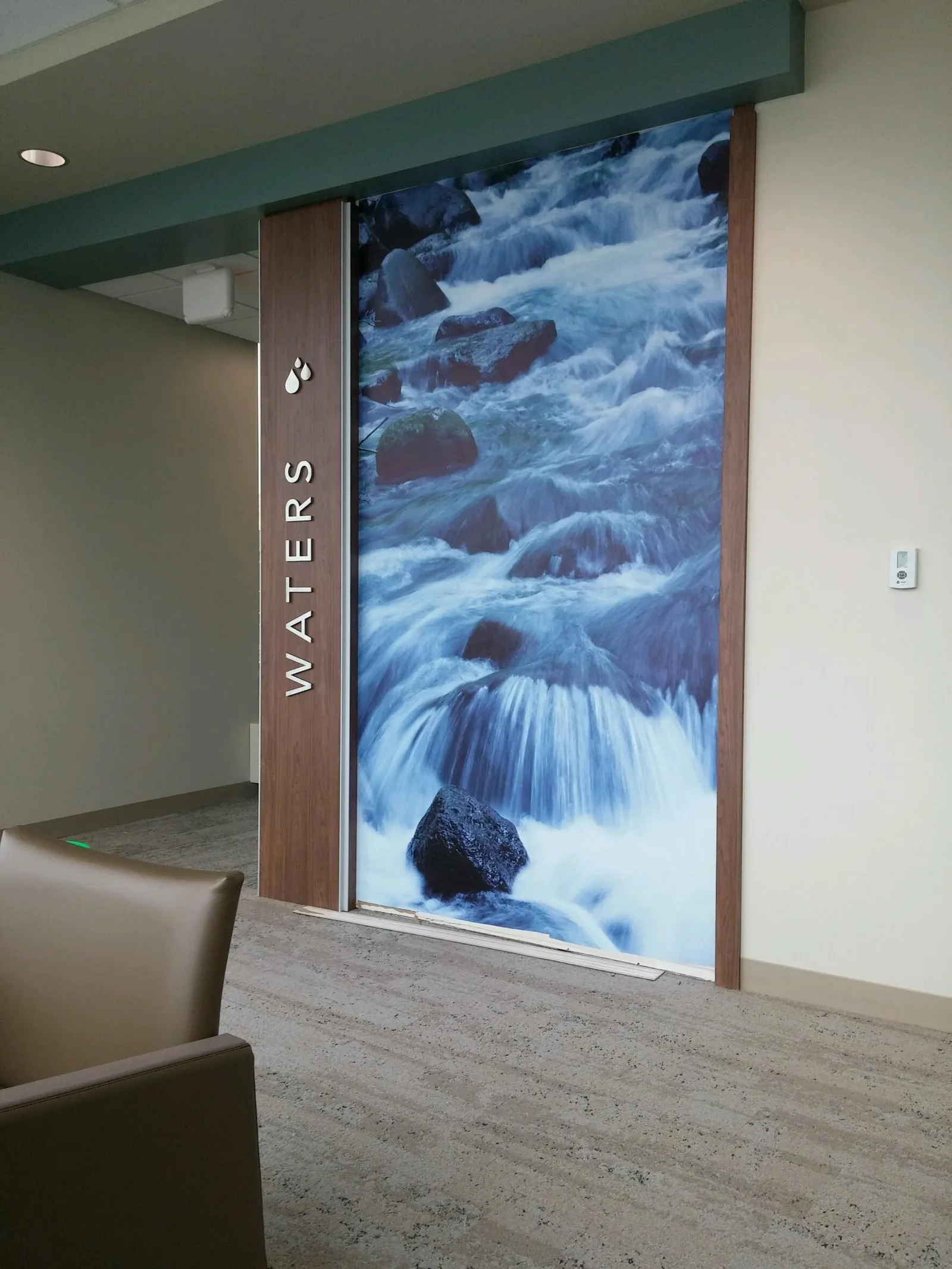 SwedishAmerican 'Waters' waiting room — floor-to-ceiling printed wall graphic of a mountain stream cascading over rocks, paired with a dimensional wood-panel 'WATERS' wordmark and water-drop icon on the entryway wall.