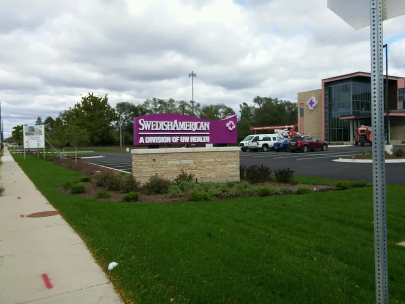 SwedishAmerican monument sign — arched purple cabinet with dimensional 'SwedishAmerican / A Division of UW Health' lettering and cross-medical-icon, mounted on a coursed-stone base with landscape shrubs, healthcare campus building with the illuminated cross behind.
