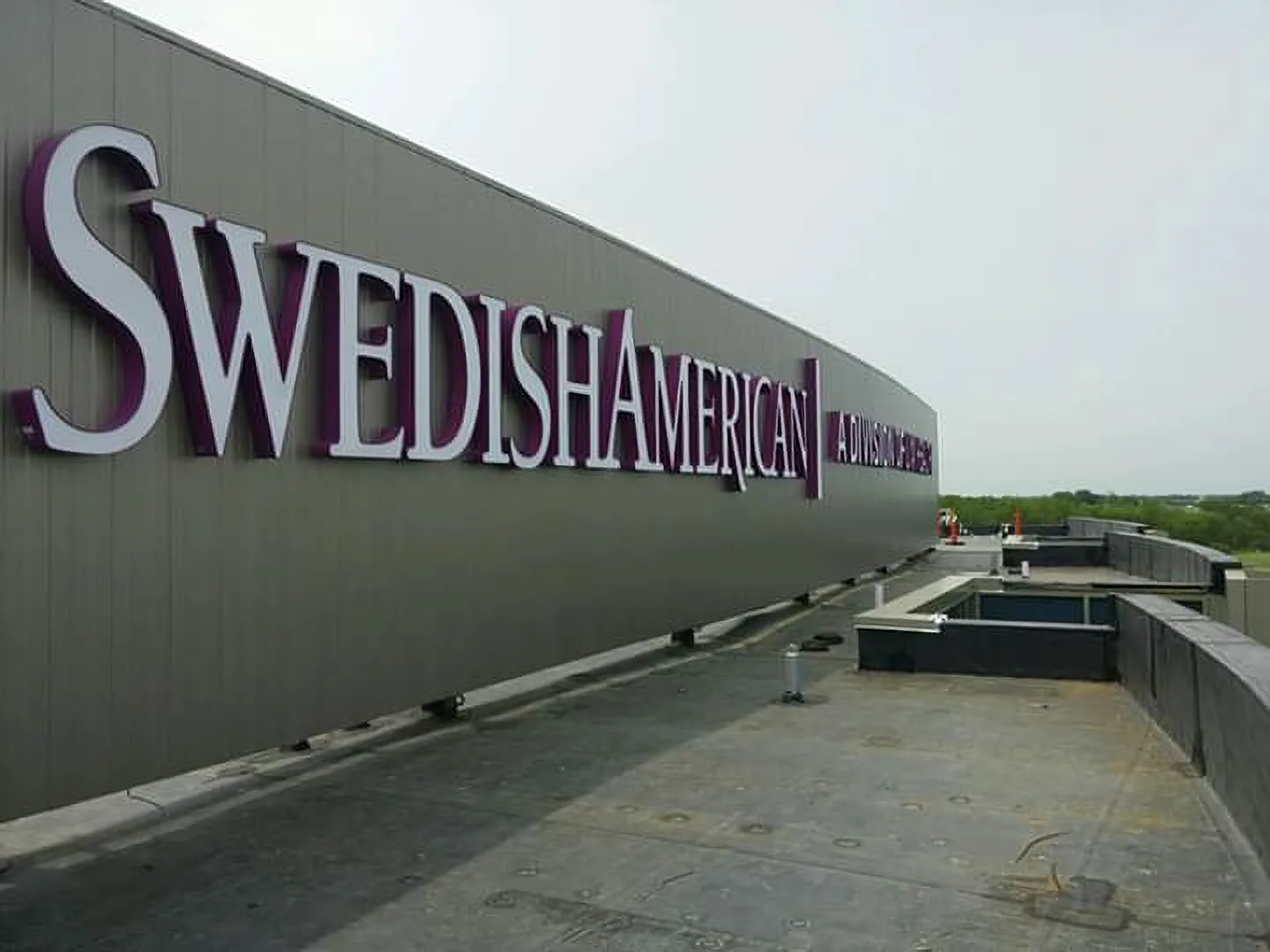SwedishAmerican rooftop building ID — architectural-scale dimensional 'SwedishAmerican / A Division of UW Health' letters in white with purple returns, mounted along a dark-panel penthouse fascia against overcast sky.