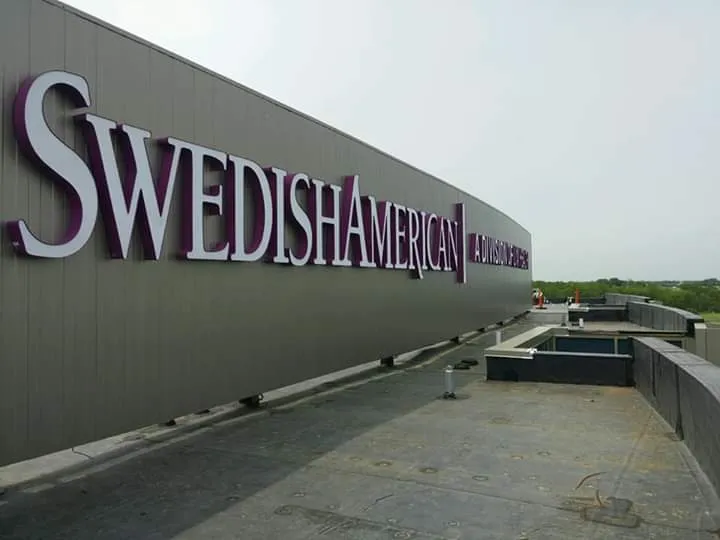 SwedishAmerican rooftop building ID — architectural-scale dimensional 'SwedishAmerican / A Division of UW Health' letters in white with purple returns, mounted along a dark-panel penthouse fascia against overcast sky.