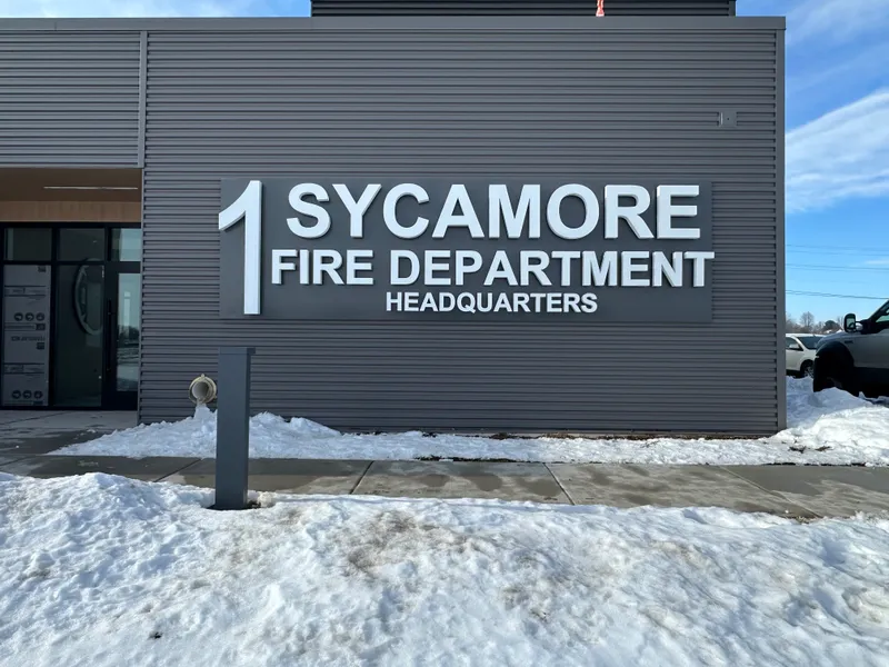 Sycamore Fire Department Headquarters building ID — clean dimensional '1 SYCAMORE FIRE DEPARTMENT HEADQUARTERS' white letters mounted on a modern dark-gray metal-siding fascia, snow on the ground, winter sun.