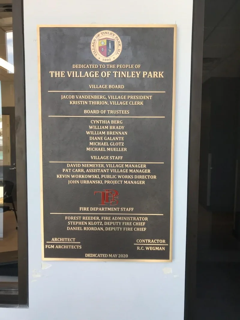Tinley Park Fire Station dedication plaque — large bronze panel with raised gold lettering listing Village Board, Trustees, and Fire Department staff, with cast village seal at top.