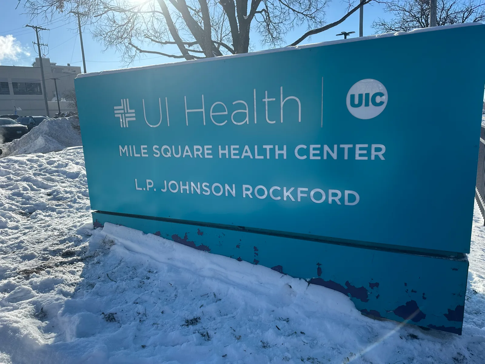 UI Health Mile Square Health Center monument sign — teal cabinet with white UIC logo and 'L.P. Johnson Rockford' lettering, photographed at three-quarter angle with winter sun flaring behind bare trees.