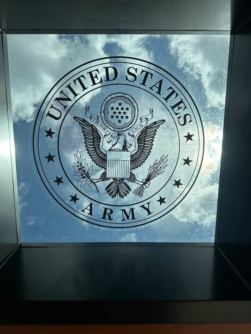 United States Army interior architectural glass — vinyl Great Seal of the United States (eagle, shield, olive branch, arrows, E PLURIBUS UNUM) applied to a mirror-finished glass panel that reflects a partly-cloudy sky, framed by a dark architectural niche.