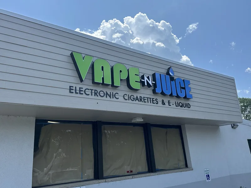 Vape-N-Juice storefront — oversized green-and-blue dimensional "VAPE-N-JUICE" letters with a water-drop accent and "Electronic Cigarettes & E-Liquid" subline on gray horizontal siding.