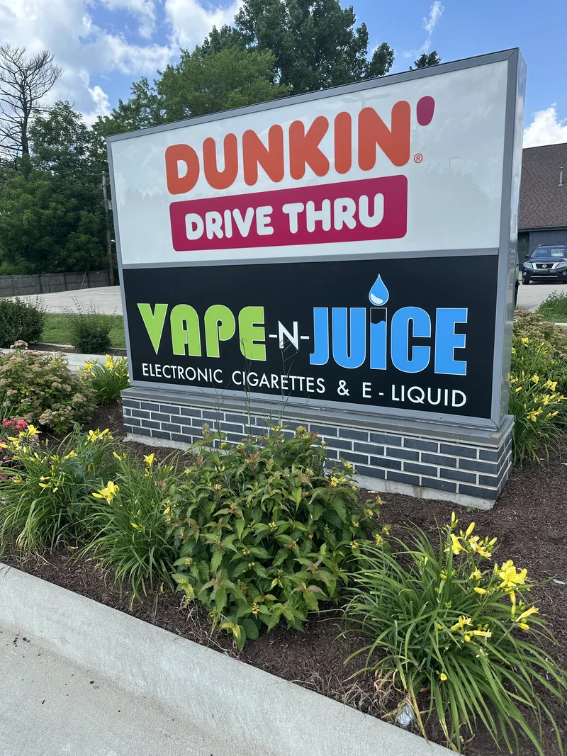 Two-tenant roadside pylon — illuminated Dunkin' Drive Thru cabinet above a black Vape-N-Juice panel with green-and-blue lettering, on a brick base in a flowering landscape bed.