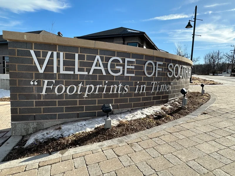 Village of South Elgin entrance monument — curved brick wall with dimensional metal letters reading 'VILLAGE OF SOUTH ELGIN' and a 'Footprints in Time' tagline, with a sculptural figure visible above the wall.