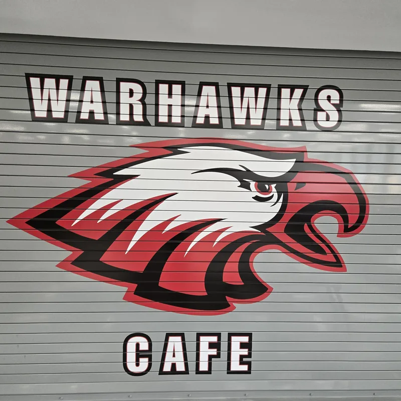 North Chicago SD187 "WARHAWKS CAFE" graphic — red, white and black Warhawk mascot printed across a ribbed metal security grille.