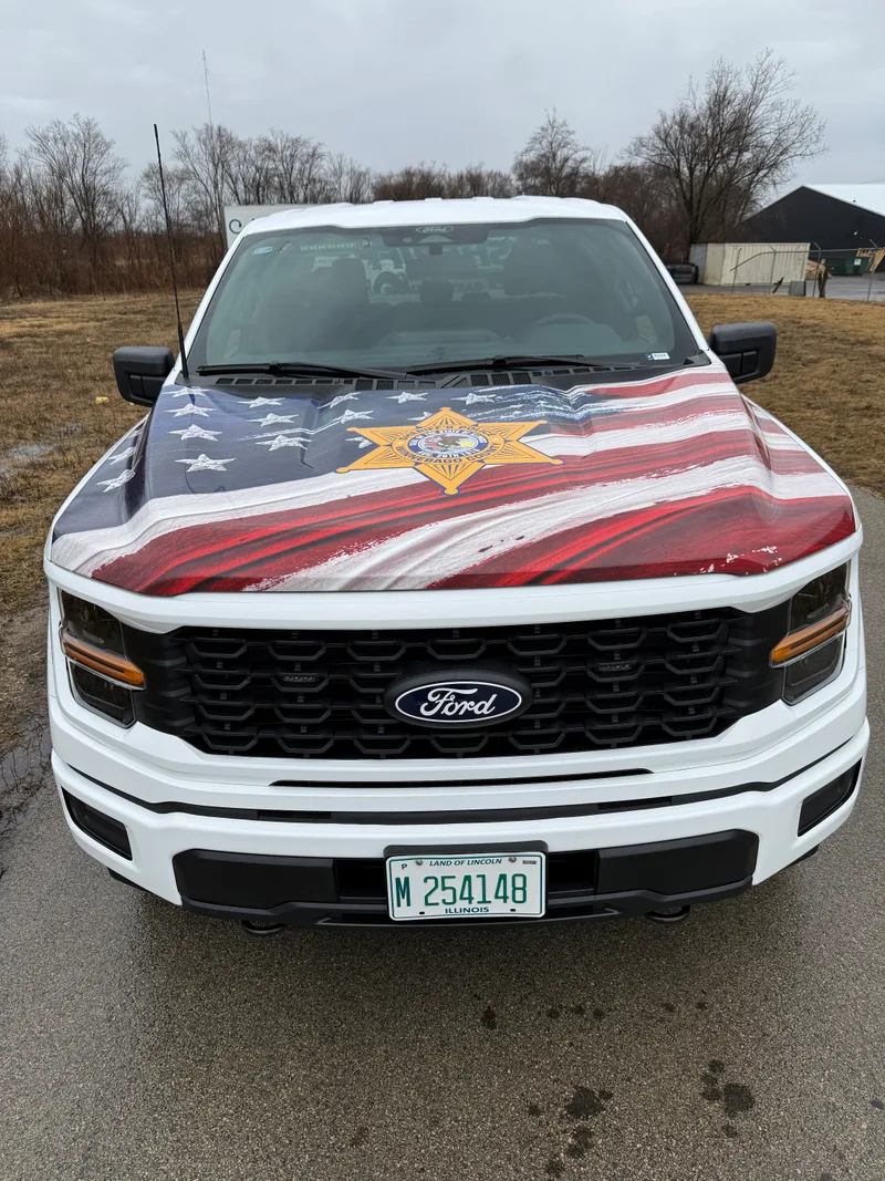 Winnebago County Sheriff Community Engagement Ford F-150 — full front-end American flag wrap with the six-point Sheriff's Police star centered on the hood, photographed after install in a rural winter setting.