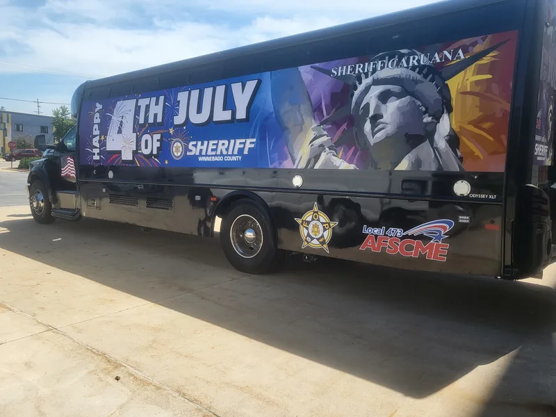 Winnebago County Sheriff transport coach outdoors — full-side "Happy 4th of July" wrap with Statue of Liberty and fireworks art on a black body.