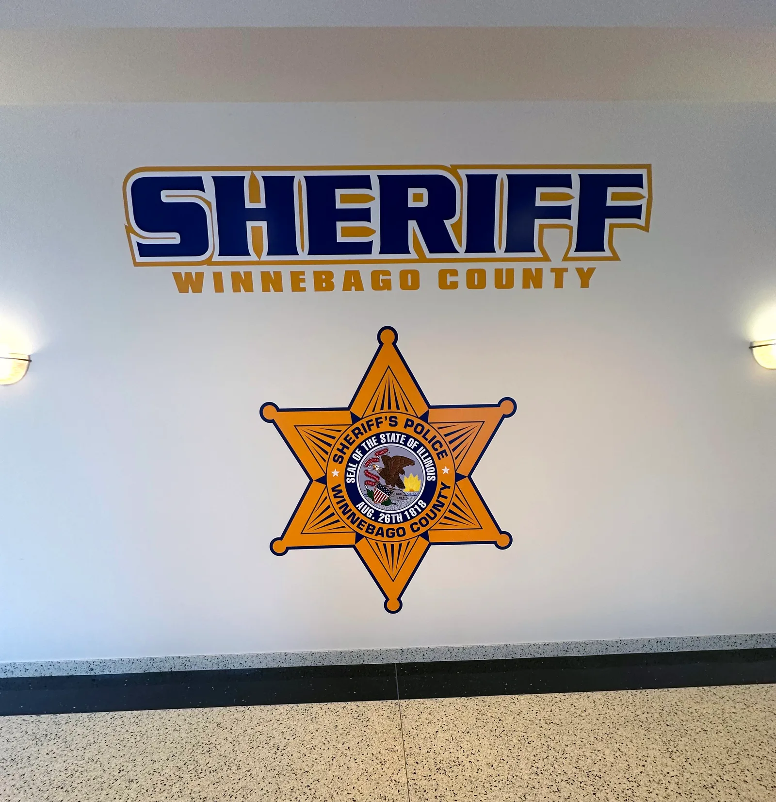 Winnebago County Sheriff's Department lobby wall — dimensional gold-and-blue 'SHERIFF / WINNEBAGO COUNTY' wordmark above the six-point Sheriff's Police star seal, mounted on a clean white wall with terrazzo floor below.