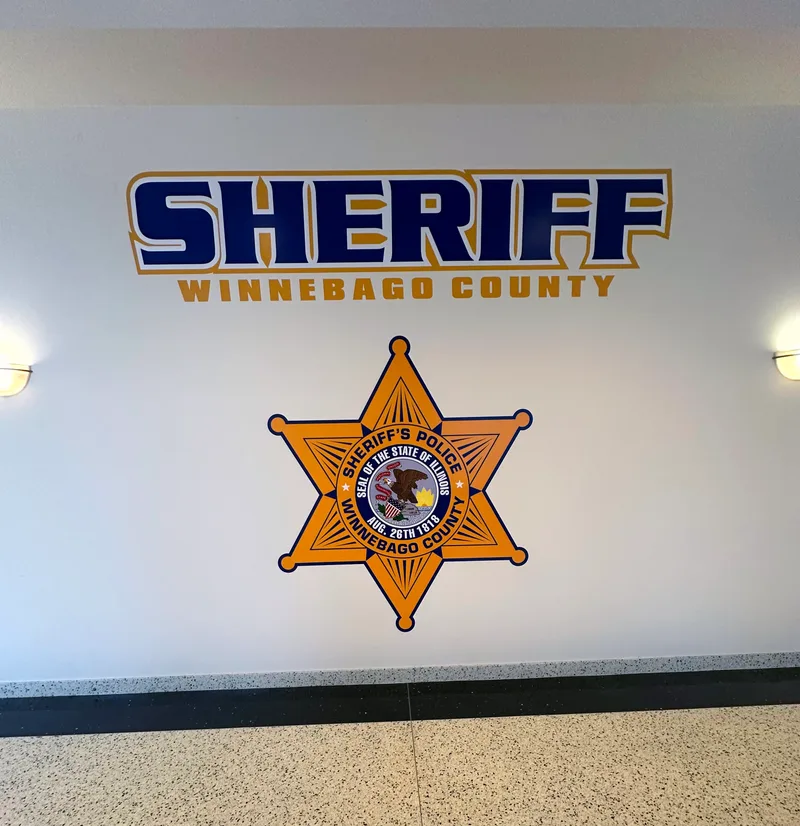 Winnebago County Sheriff's Department lobby wall — dimensional gold-and-blue 'SHERIFF / WINNEBAGO COUNTY' wordmark above the six-point Sheriff's Police star seal, mounted on a clean white wall with terrazzo floor below.