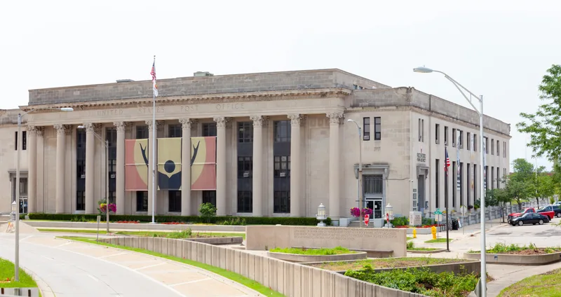 Rockford Park District banners at the Webbs Norman Center (former US Post Office) — three large cream, maroon, and gold building banners featuring the Park District's raised-arms figure emblem, hung between the historic Corinthian columns of the neoclassical civic building, with the American flag and the Rockford founders' dedication plaque in view.