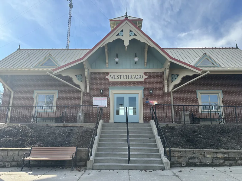 Restored West Chicago train station — "WEST CHICAGO" dimensional letters on an oval panel beneath the Victorian gable trim, above the brick entry stairs.